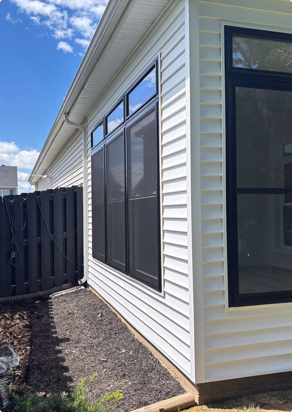 White-sided house with large black-framed windows overlooks a freshly cleared garden bed beside a dark fence under a bright blue sky, creating a calm, sunlit scene.