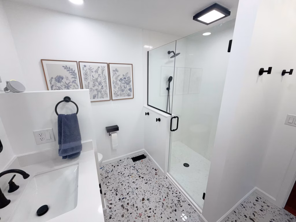 Bright modern bathroom with a glass walk-in shower, black fixtures, white vanity, terrazzo floor, and framed botanical prints; clean, calm, minimalist feel.