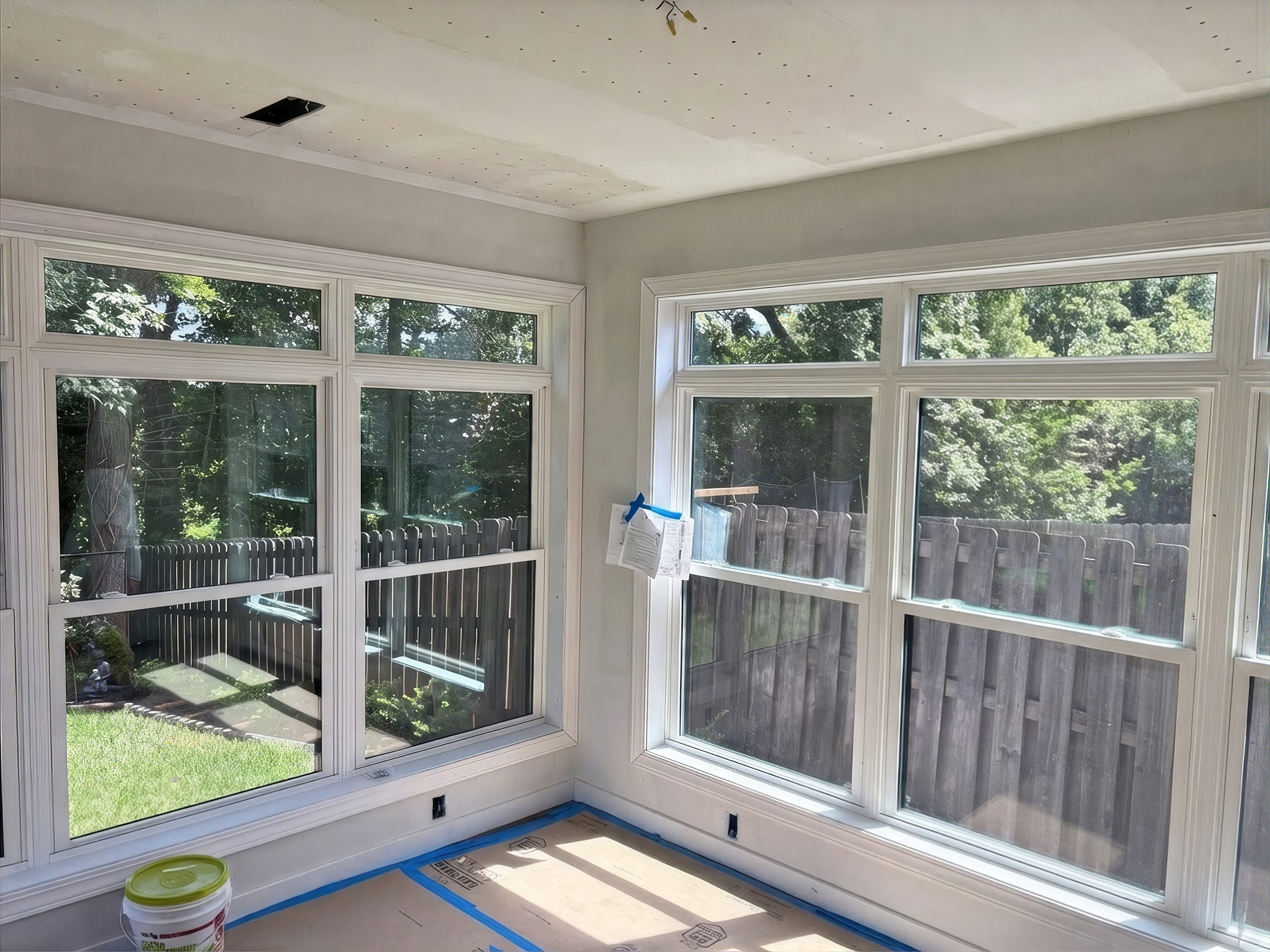 Sunlit enclosed porch with large white-framed windows overlooking a green yard and wooden fence; blue painter’s tape and a paint bucket suggest renovation.
