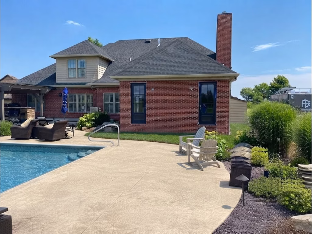 Sunny backyard with a red-brick house, swimming pool, and spacious concrete patio furnished with chairs; green landscaping creates a calm, inviting outdoor retreat.