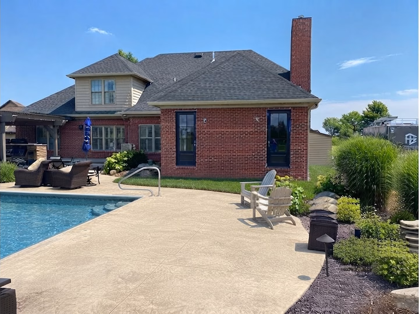 Sunny backyard with a red-brick house, swimming pool, and spacious concrete patio furnished with chairs; green landscaping creates a calm, inviting outdoor retreat.