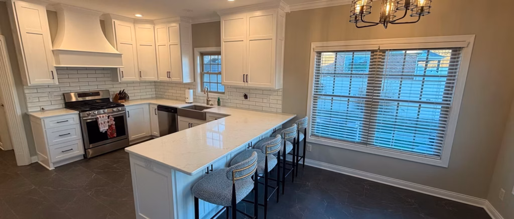 Bright modern kitchen with white cabinets, subway tile, stainless range, large island and three stools beside a sunlit window, creating a clean, welcoming feel.