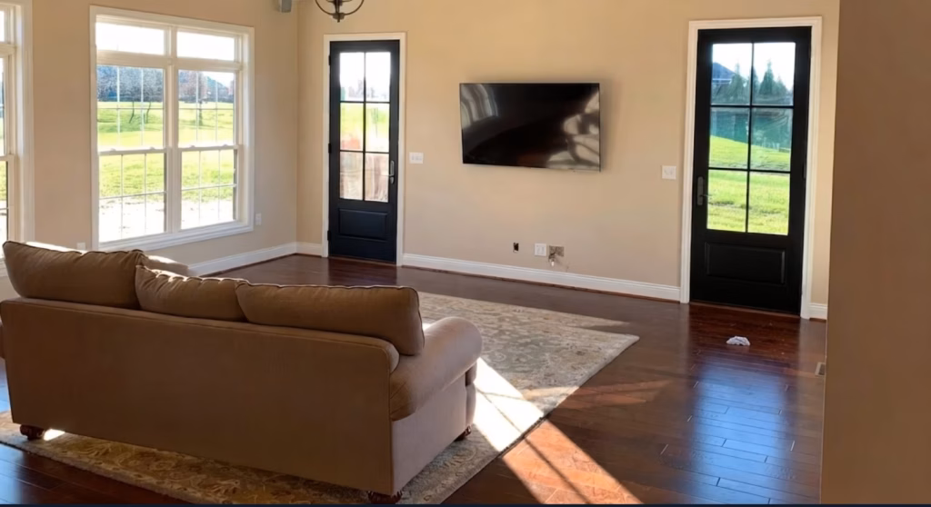 Bright living room with a brown sectional, wall-mounted TV, large windows and black glass doors; sunlight falls across hardwood floors, creating a calm feel.