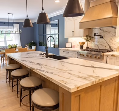 Modern blue-and-white kitchen with a marble waterfall island, four tan stools, black pendant lights, and a farmhouse sink, creating a warm, stylish feel.