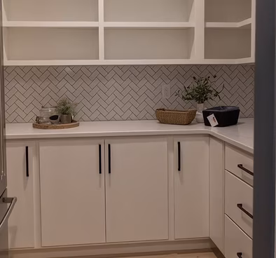A clean, modern white kitchen with open shelving, geometric herringbone tile, dark cabinet handles, and small plants and baskets on the counter; calm, minimalist tone.