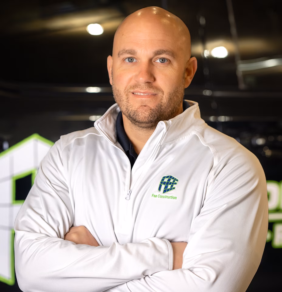Josh Fee stands with arms crossed in a white Free Cooperative jacket, smiling slightly in a dim indoor setting with green-and-white signage.