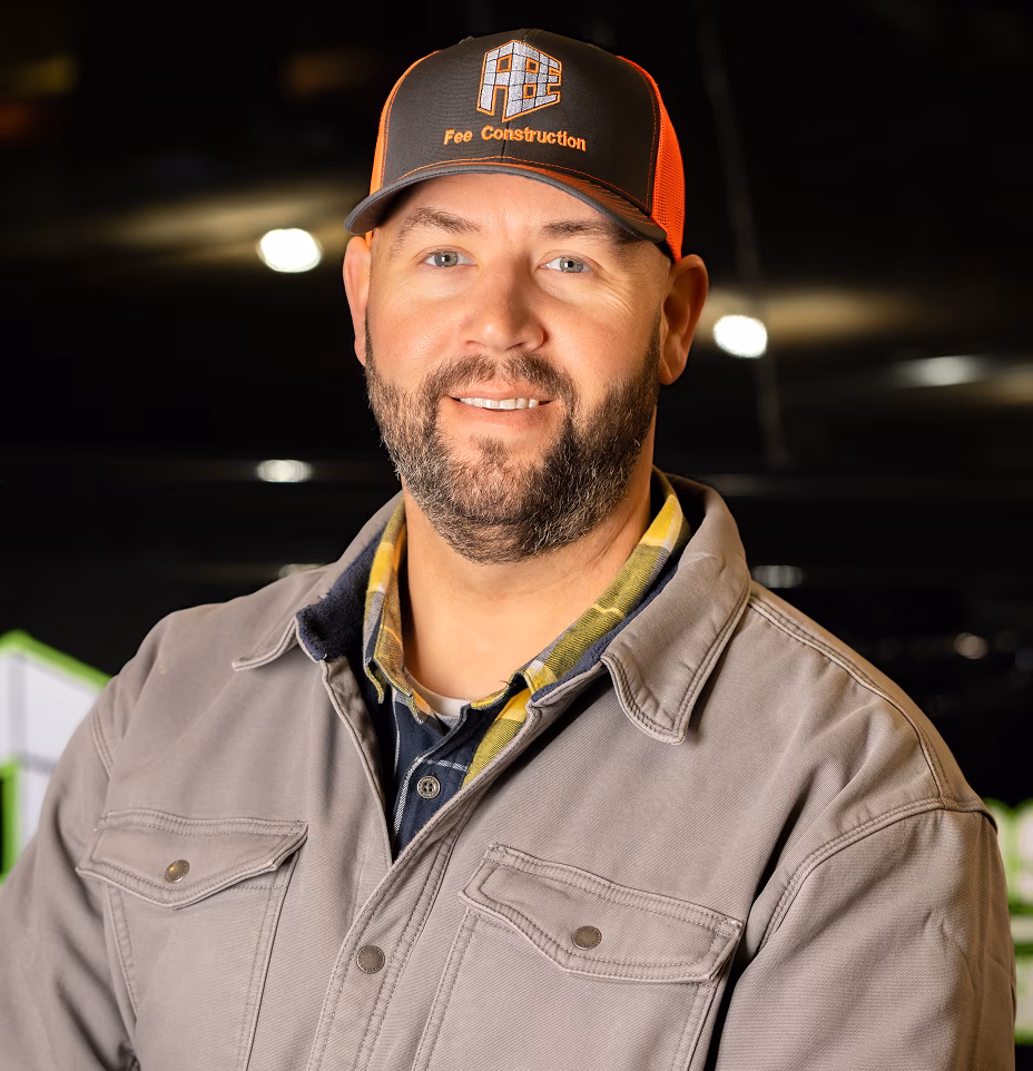 A smiling Jared fee in a gray jacket and black-and-orange Fox Construction cap stands in a dim indoor worksite, conveying a friendly, confident tone.