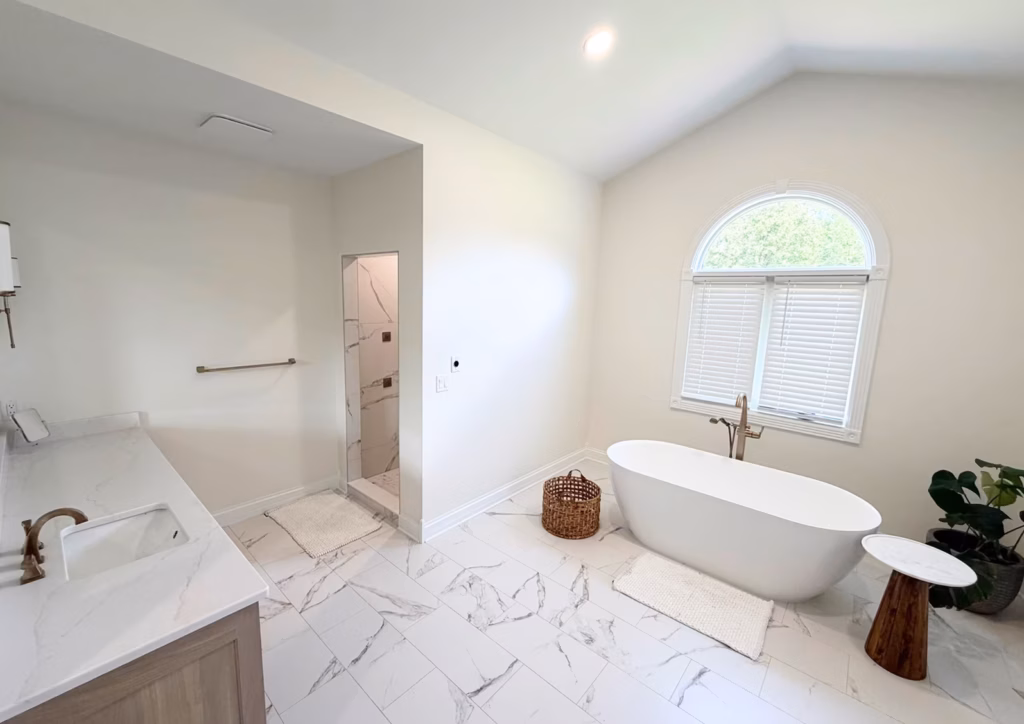 Bright, serene bathroom with a freestanding tub beneath an arched window, marble floors, a stone vanity, woven basket, and greenery.