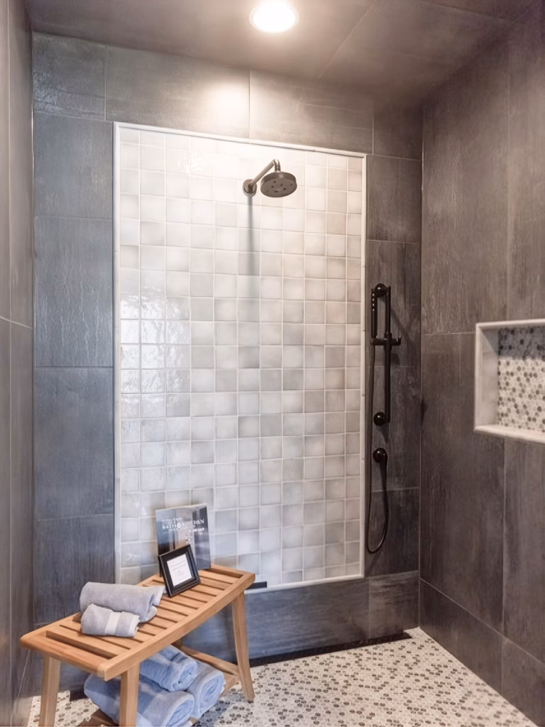Modern gray stone walk-in shower with white tile, rainfall head, handheld sprayer, recessed shelf, and teak bench stacked with towels creates a calm spa-like feel.