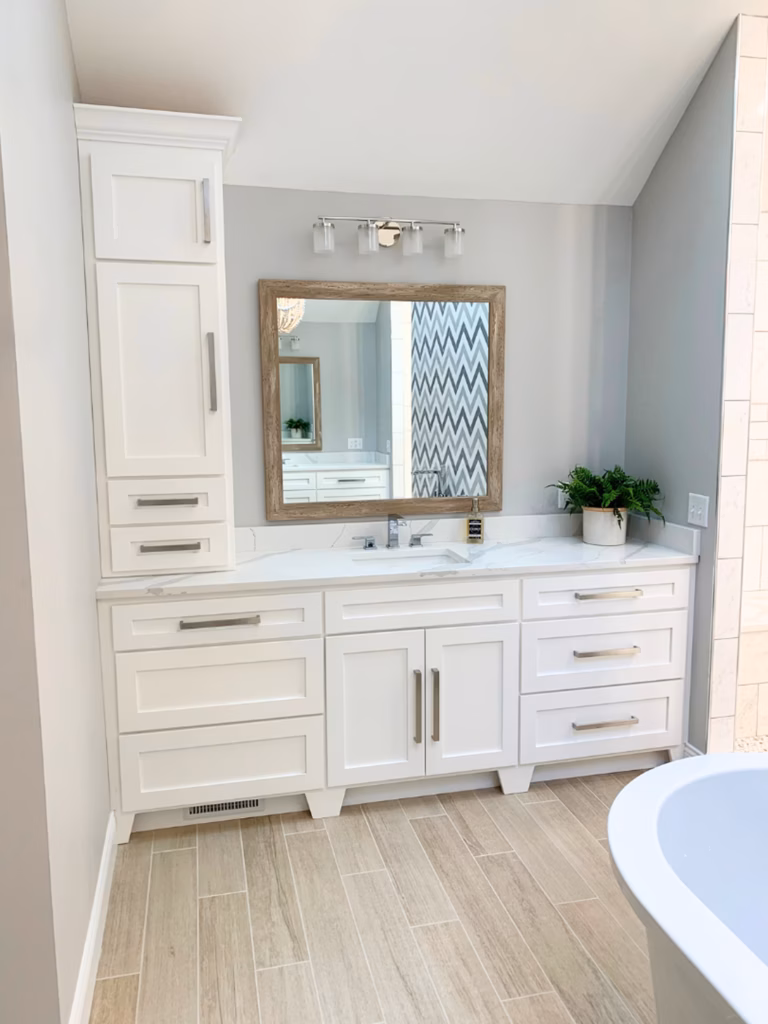 Bright, serene bathroom with a white built-in vanity, brass handles, framed mirror, small green plant, and pale wood-look tile beside a curved tub.