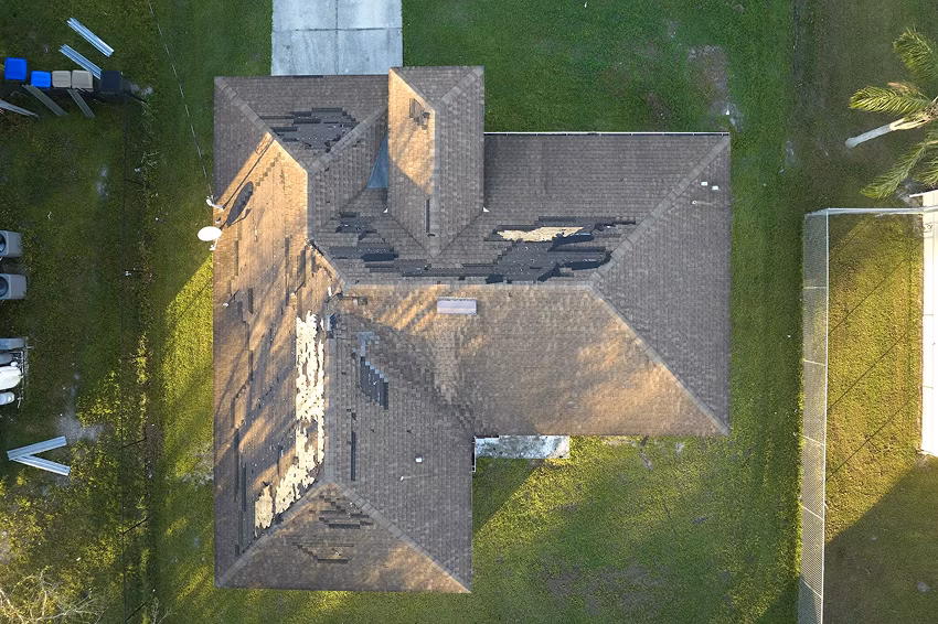 Overhead aerial view of a weathered, cross-shaped house roof surrounded by green lawn, with long afternoon shadows creating a quiet, rural mood.
