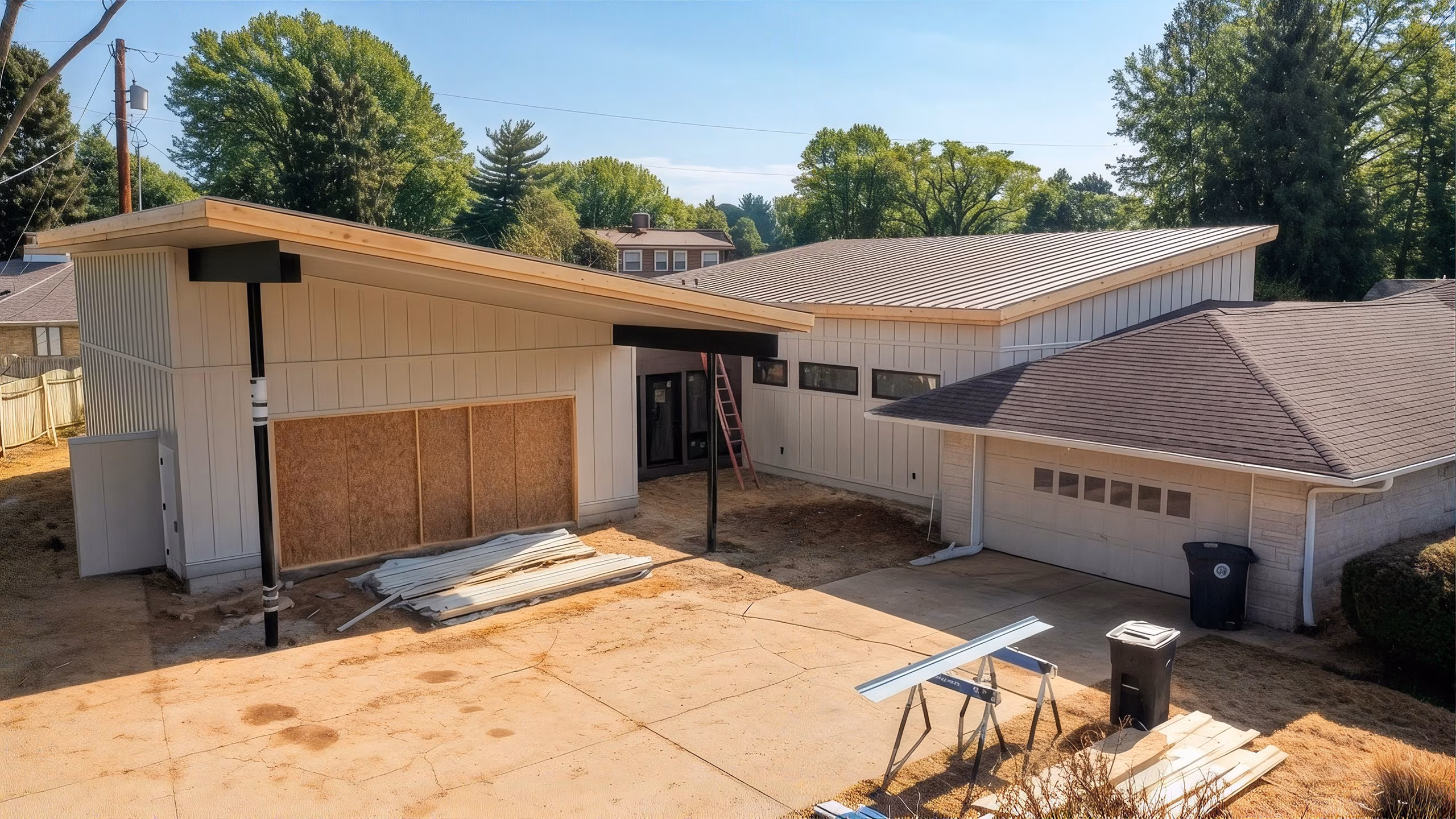 A partially renovated suburban garage with plywood-covered openings, construction materials, a ladder, and sawhorses sits in bright midday sun, creating a quiet worksite scene.