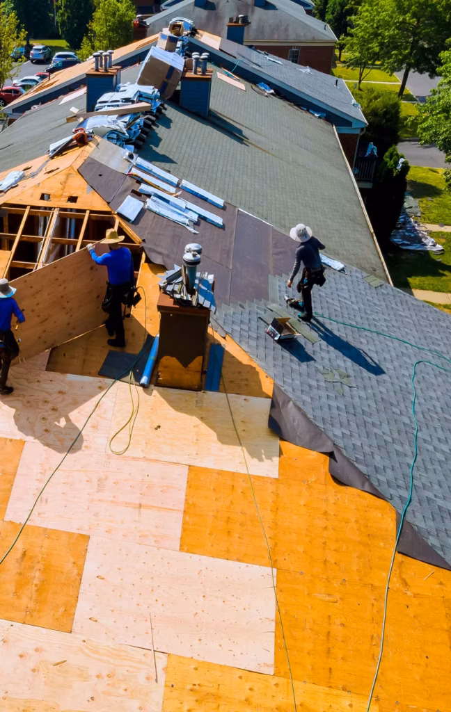 Aerial view of roofers repairing a house, installing gray shingles beside broad areas of exposed plywood in a sunny, industrious scene.