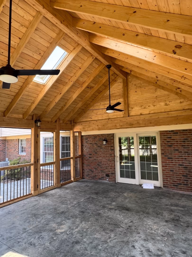 An empty covered patio with a vaulted honey-colored timber ceiling, exposed beams, skylight, ceiling fans, brick walls, glass doors, and a concrete floor. The space feels bright and spacious.