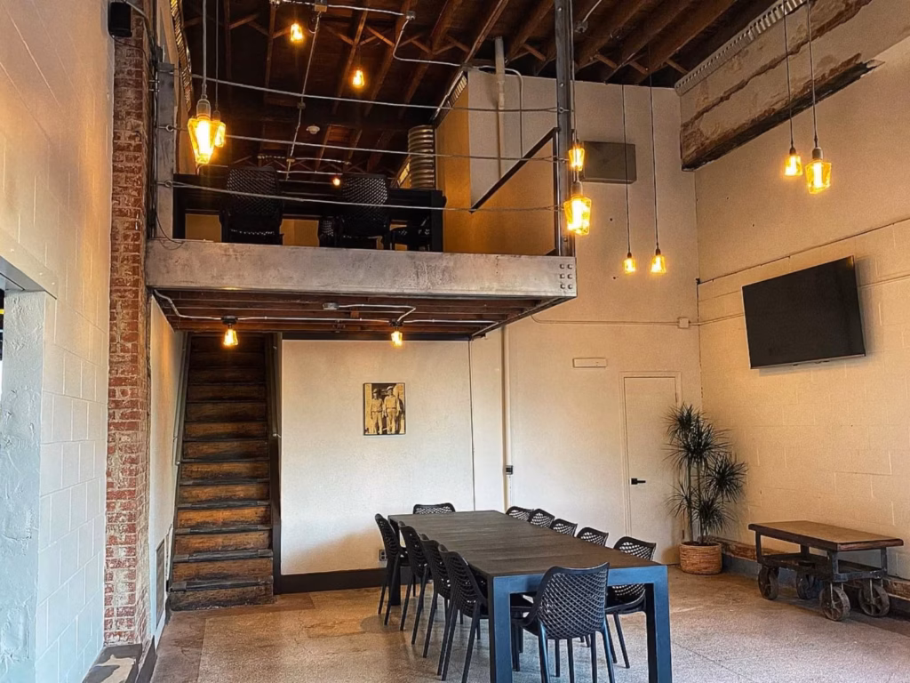 Rustic industrial dining room with a long table and black chairs, wooden stairs to a mezzanine, exposed beams, and warm pendant lights.