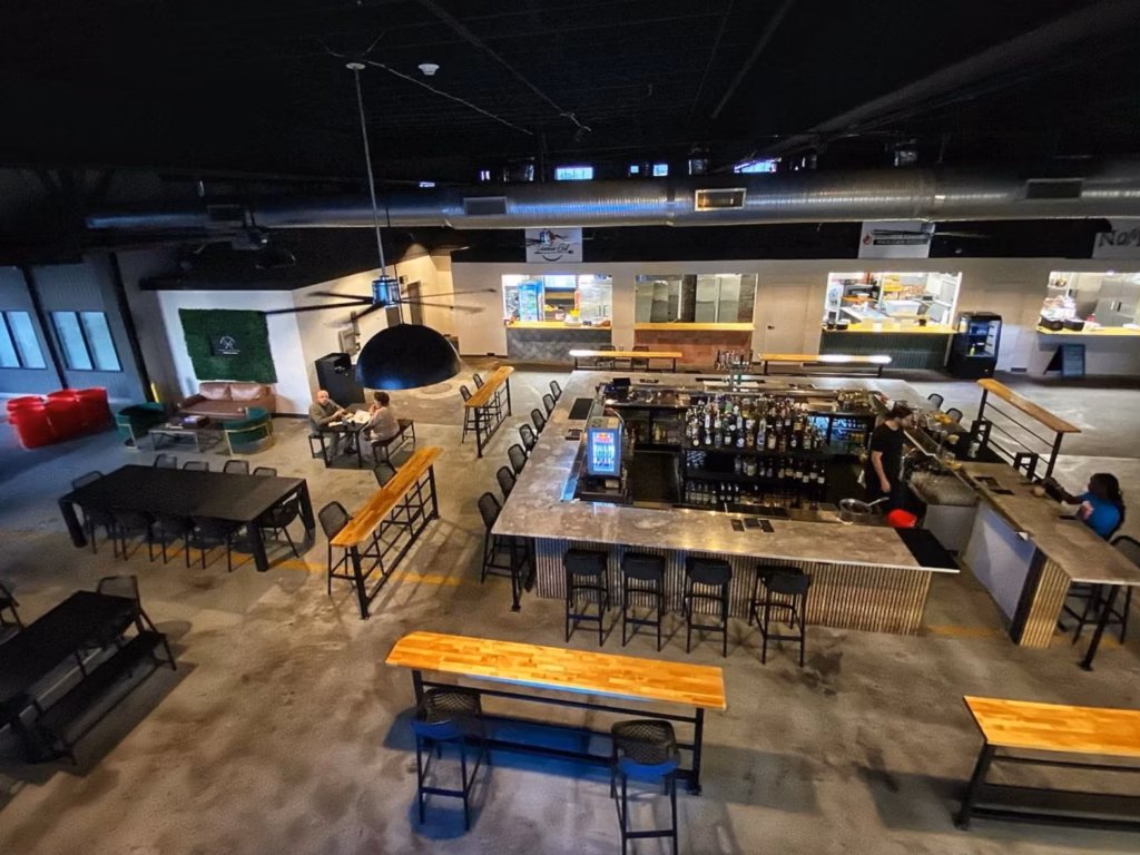 An elevated view of a modern bar and restaurant with a long counter, rows of stools, wooden tables, hanging lights, and a few patrons; the atmosphere feels relaxed and industrial.