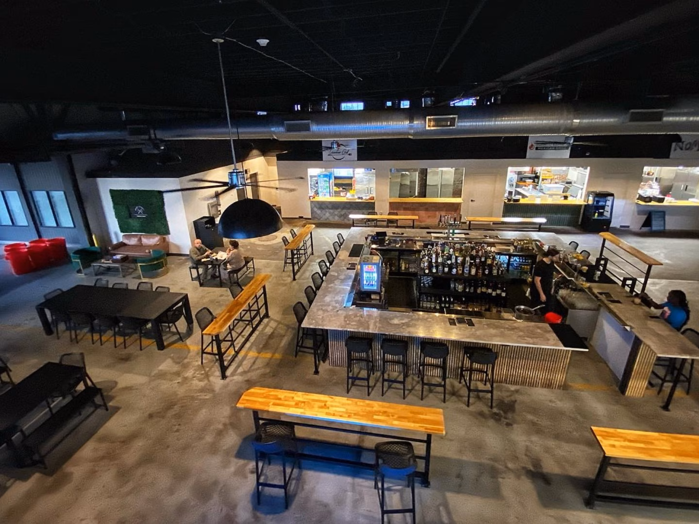 An elevated view of a modern bar and restaurant with a long counter, rows of stools, wooden tables, hanging lights, and a few patrons; the atmosphere feels relaxed and industrial.