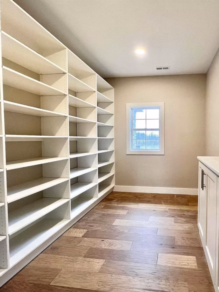 A bright, empty walk-in pantry with floor-to-ceiling white shelves, a small window, warm wood flooring, and a clean, orderly feel.