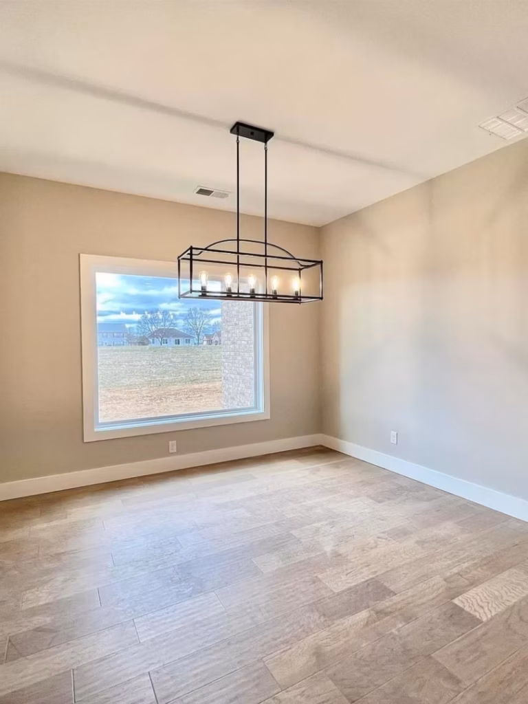Bright, empty dining room with beige walls, wood-look flooring, a large window overlooking homes, and a black cage-style pendant light; calm, neutral interior.