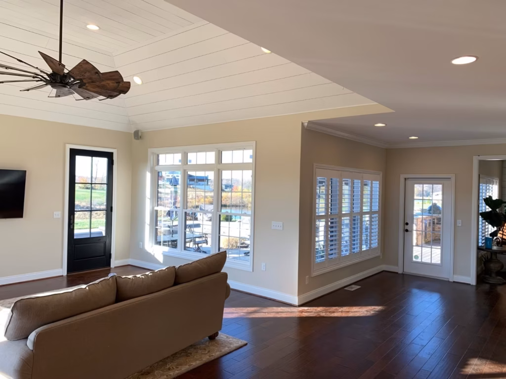Sunlit open-plan living room with a brown sofa, dark wood floors, large windows, and a ceiling fan; bright, spacious, and welcoming.