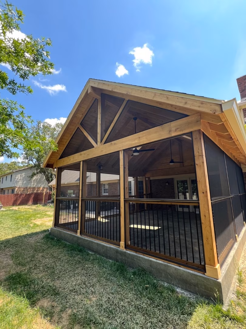 A sunny outdoor view of a covered wooden patio with exposed timber beams, black railings, and shaded seating beside a grassy yard.