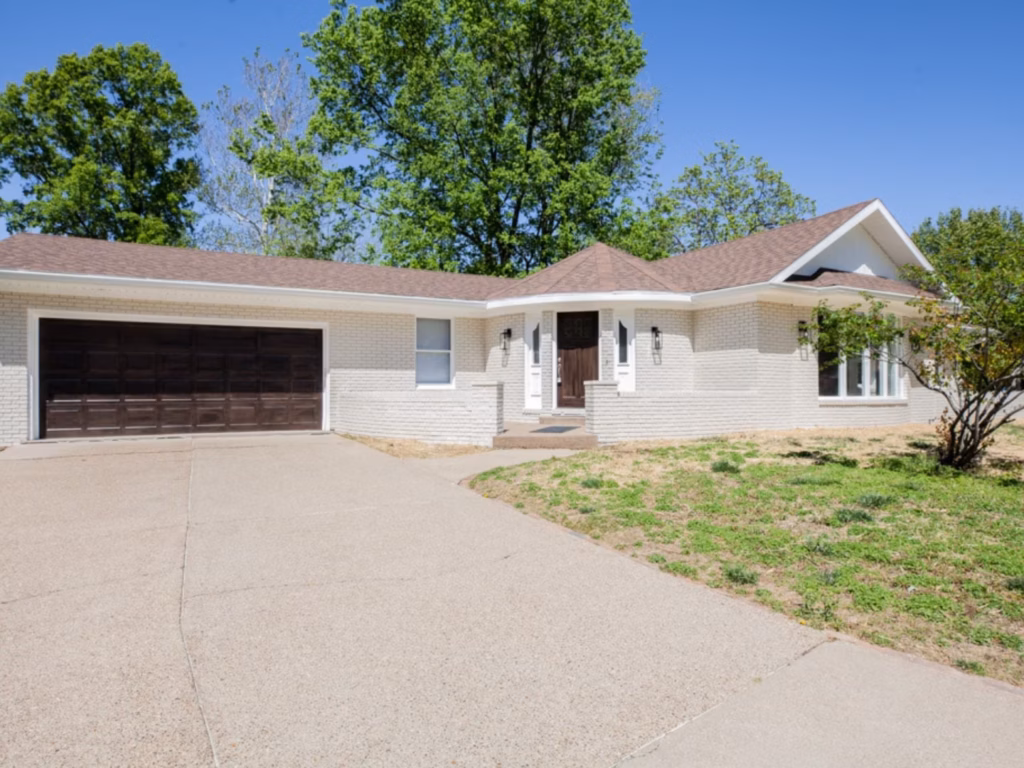 A single-story white brick suburban home with a brown roof, dark garage door, wide driveway, and patchy lawn sits beneath leafy trees in bright sunshine.