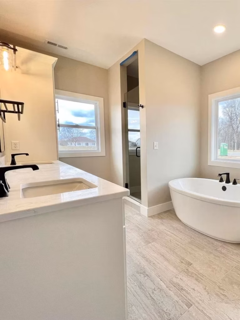 Bright modern bathroom with a white double-sink vanity, glass shower, and freestanding tub beside two windows; the clean, neutral space feels calm and airy.