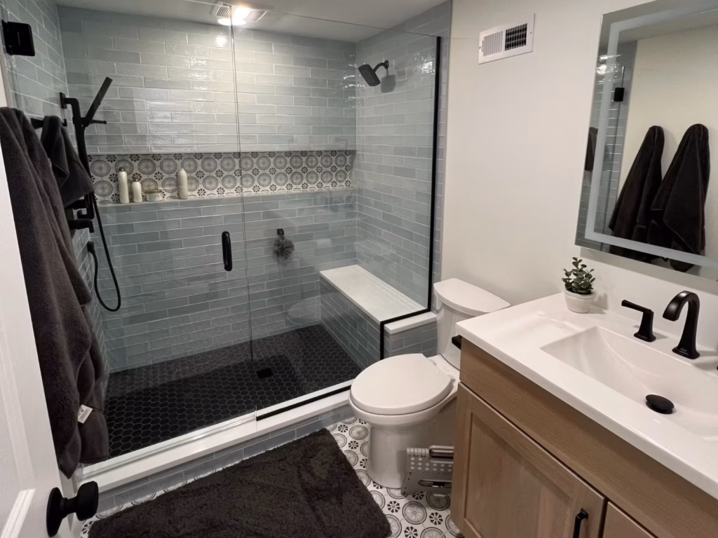 Modern bathroom with a spacious glass-enclosed shower, pale blue tile, patterned accent strip, white toilet and sink, and dark towels; clean, calm feel.
