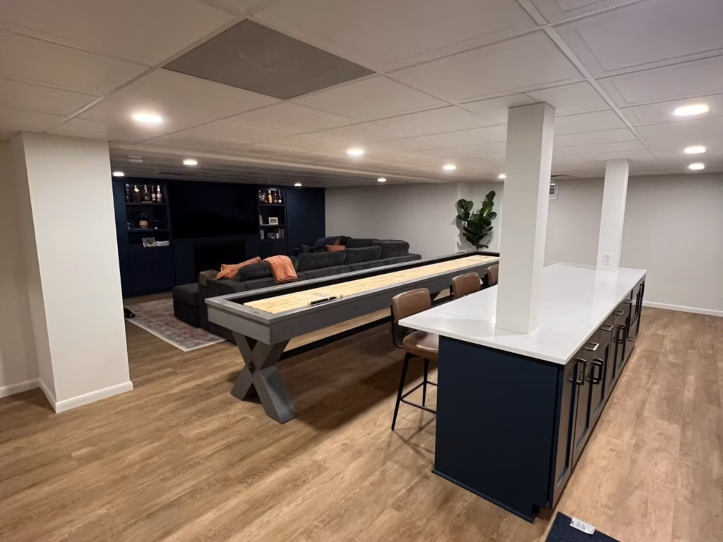 Modern finished basement with a long shuffleboard table, dark lounge seating, built-in shelving, and a white kitchen island with stools; warm lighting creates a relaxed, inviting atmosphere.