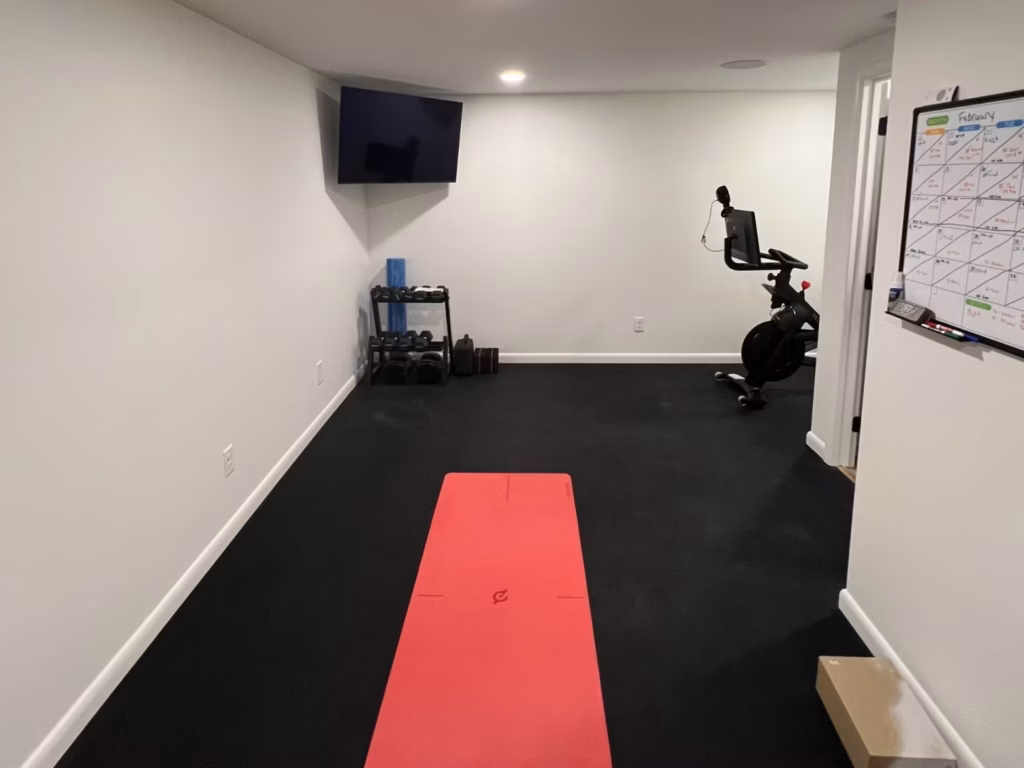 A quiet home gym with a red exercise mat on black flooring, dumbbells and a stationary bike, plus a wall-mounted TV and workout calendar.