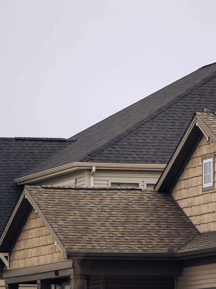 Close-up of a suburban home’s intersecting gray shingle roofs and beige siding beneath an overcast sky, creating a quiet, neutral mood.